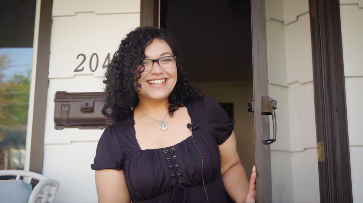 a dark haired young woman opening her front door and smiling at the camera